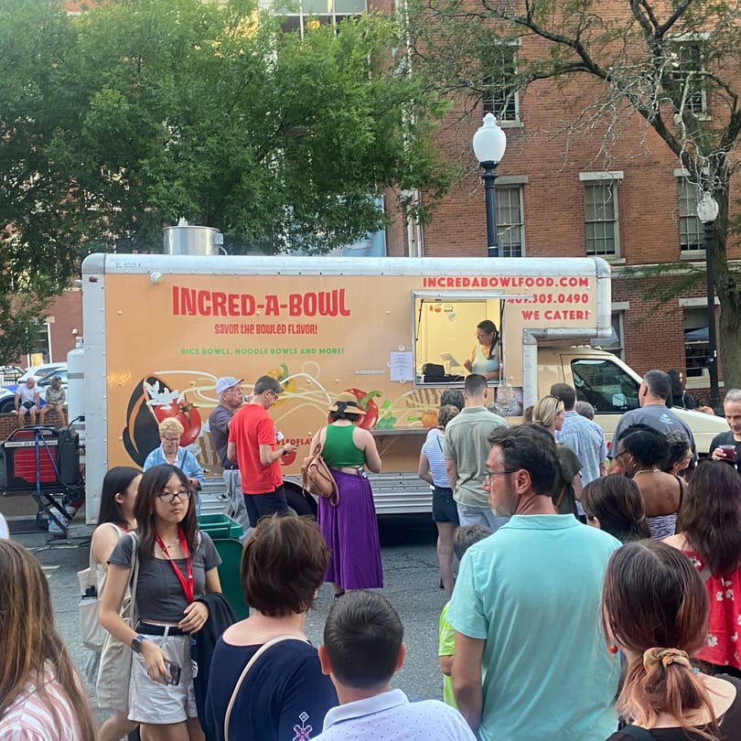 A crowd of people gathers around the Incred-A-Bowl Food Co. food truck on a city street.