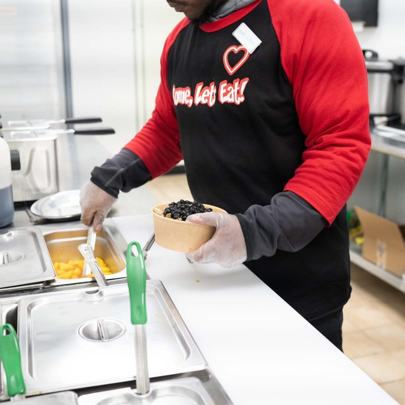 A person in a red and black shirt serving black beans into a bowl at Incred-A-Bowl Food Co.
