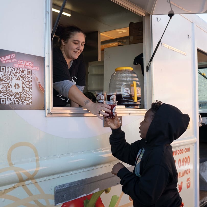 A smiling Incred-A-Bowl Food Co. worker hands a drink to a child from a food truck window.