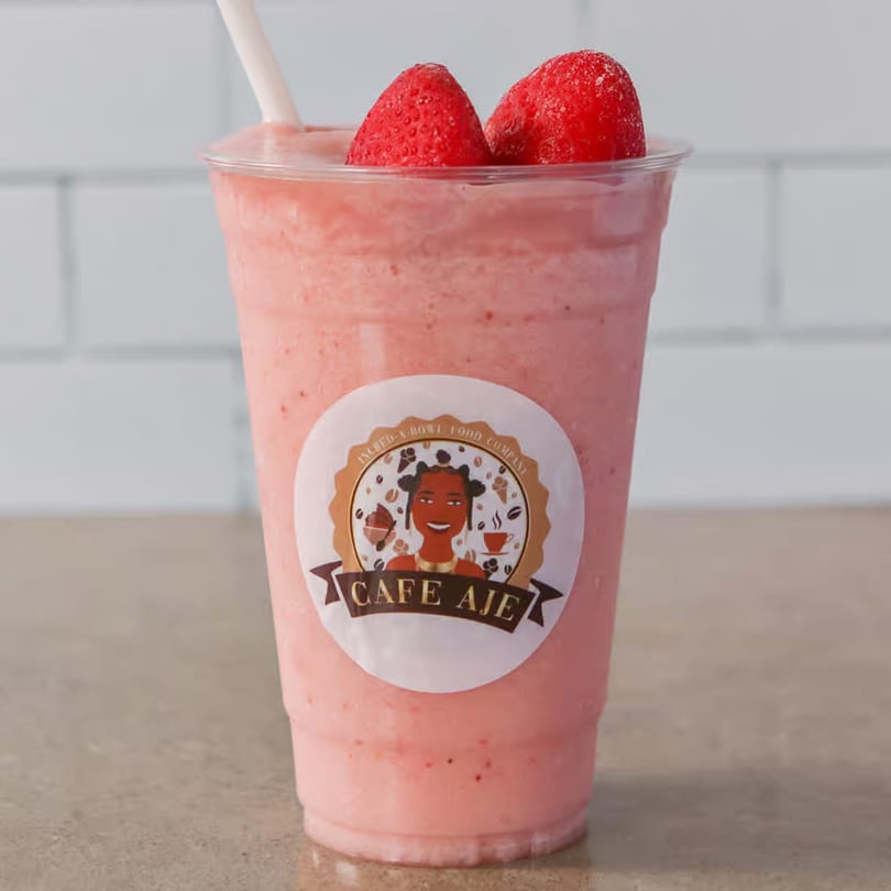 A strawberry smoothie in a clear plastic cup with two whole strawberries on top and a Cafe Aje logo.
