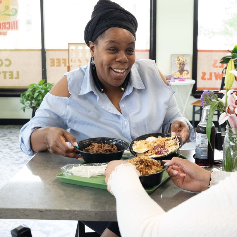 Two women at Incred-A-Bowl Food Co. enjoying healthy meals in black bowls on a gray table.